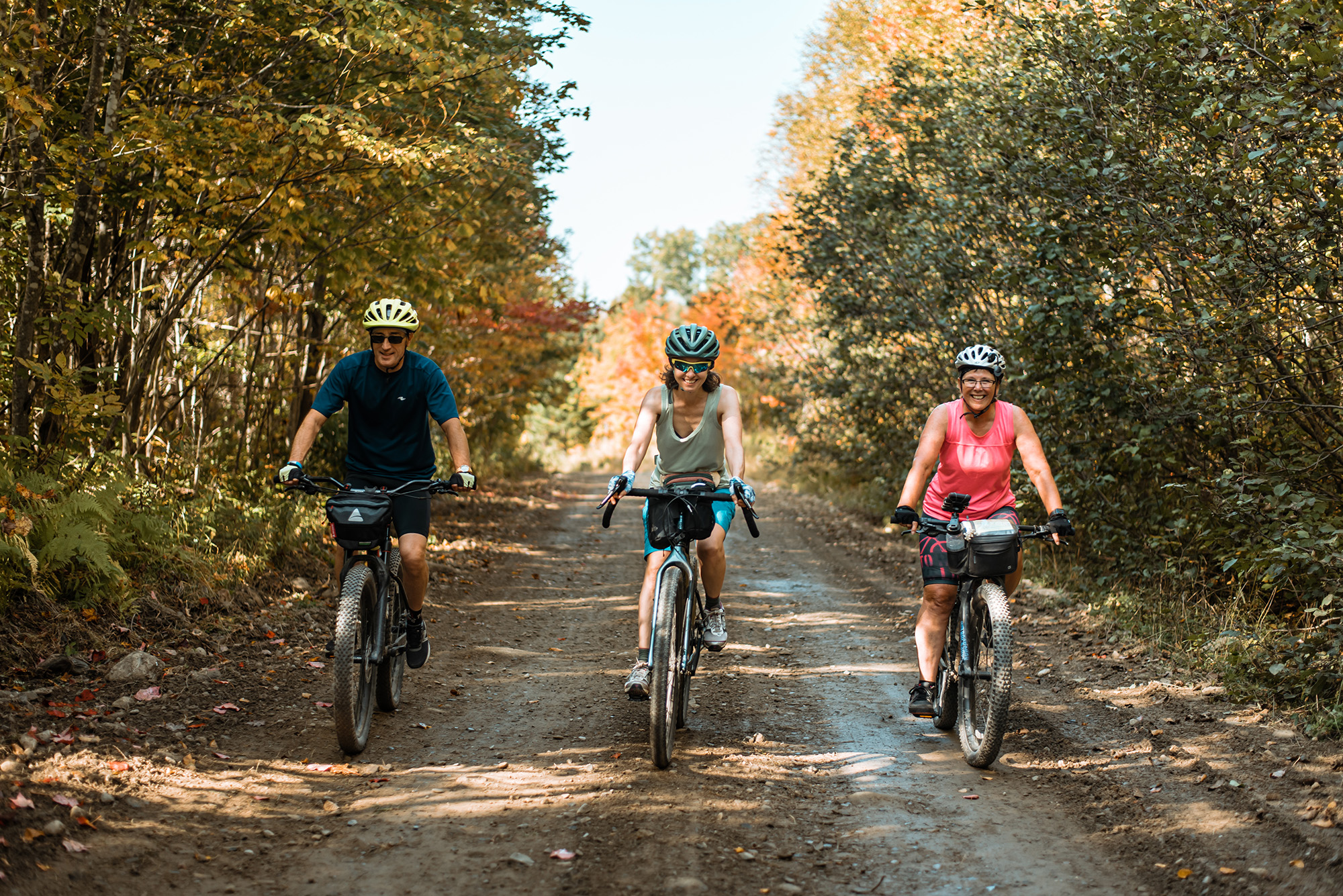 Photo by Garrick Hoffman. Three smiling people gravel biking on a wide trail in the woods.